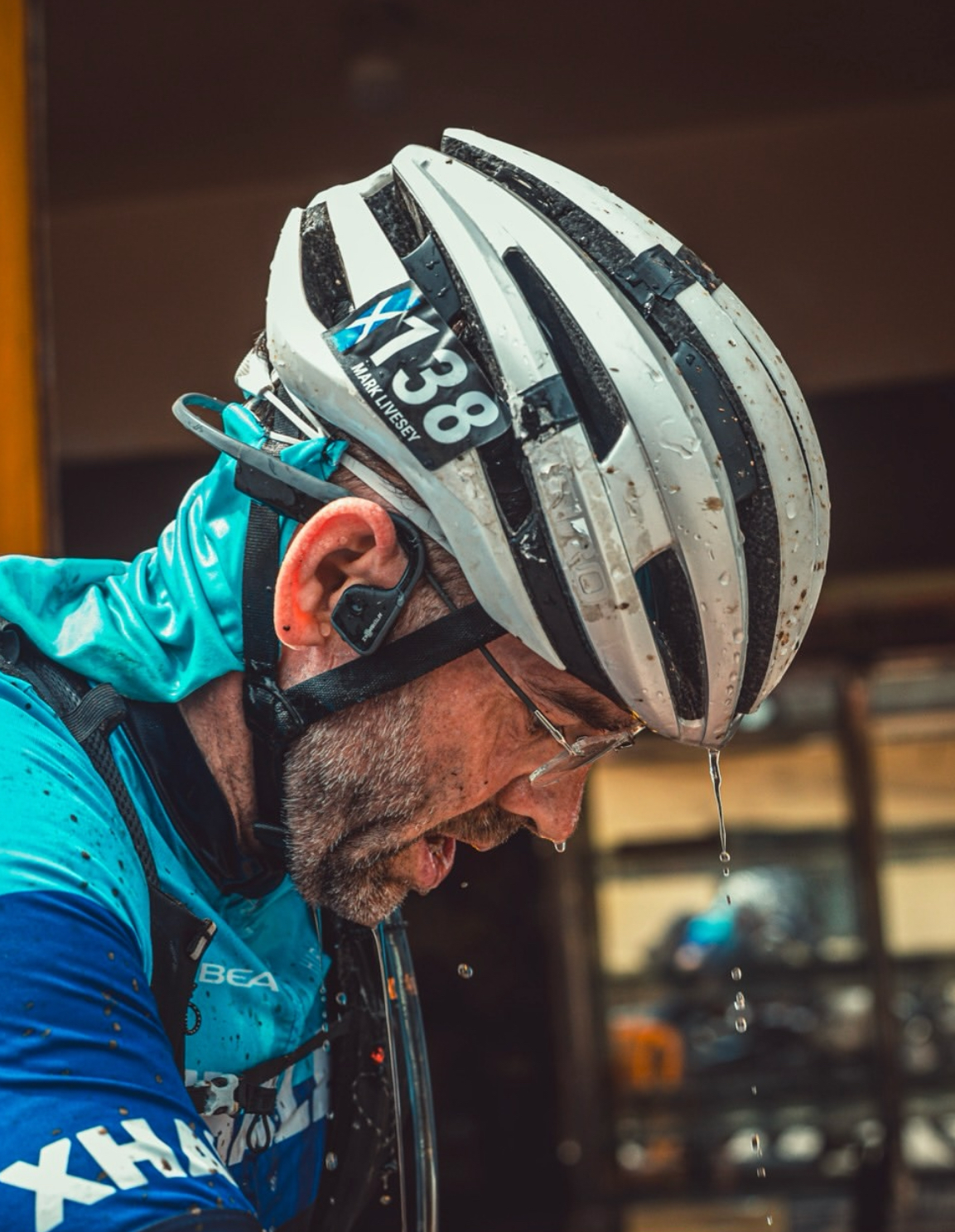 Close-up of a cyclist wearing a rain-spattered helmet and blue jersey, head bowed with water dripping