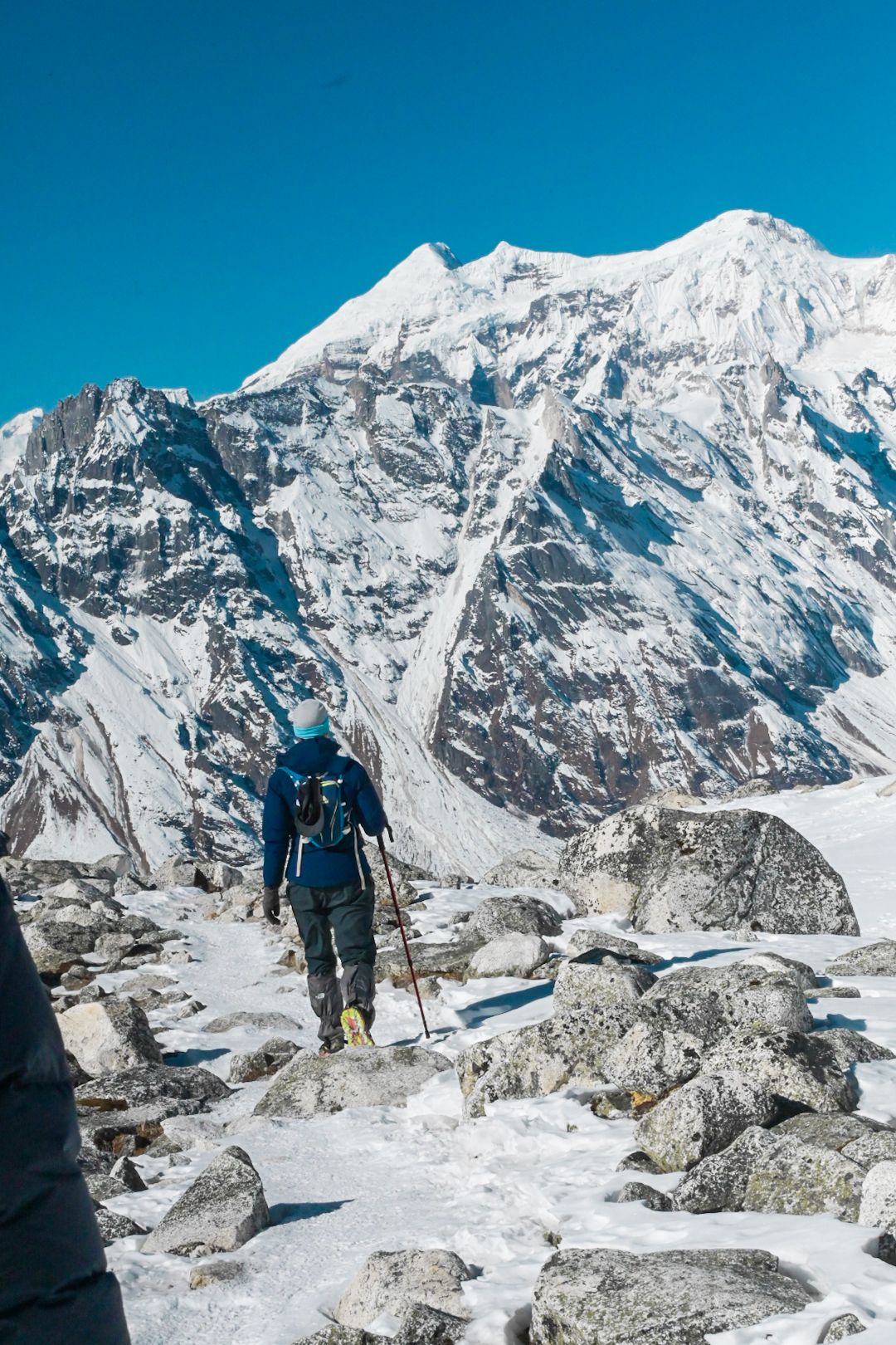 Trekker crossing a snowy boulder field beneath a huge Himalayan face