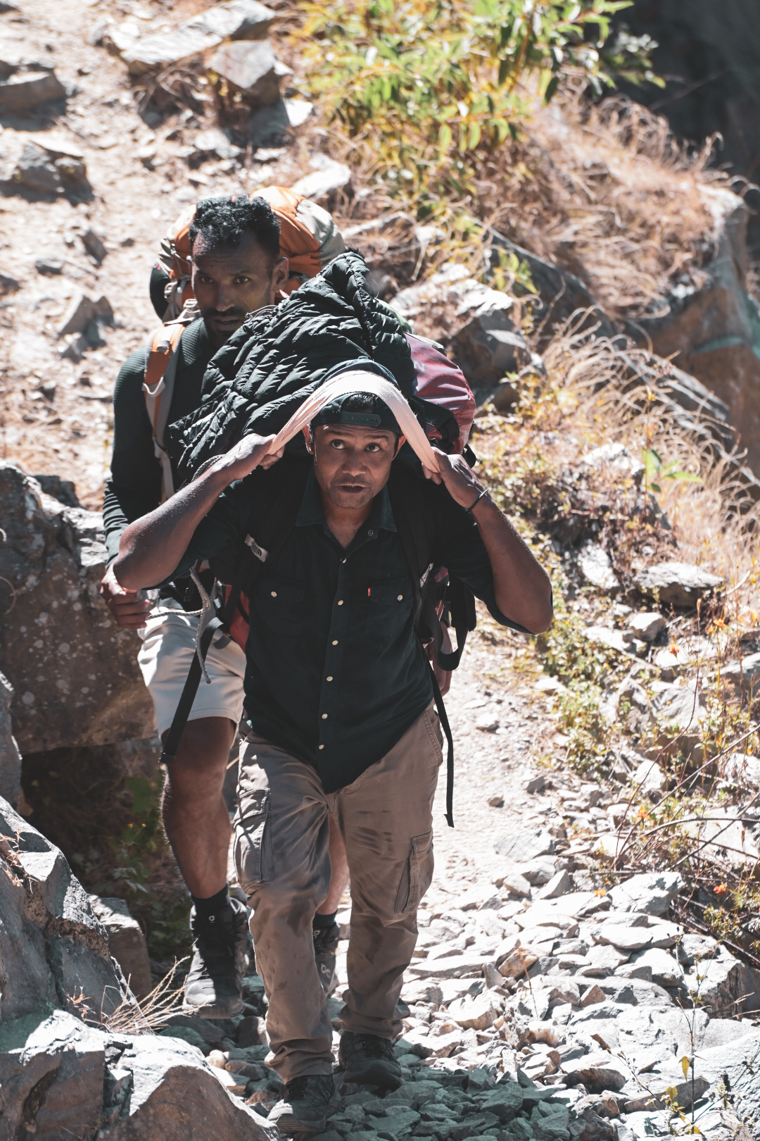 Two porters carrying loaded packs up a rocky mountain trail
