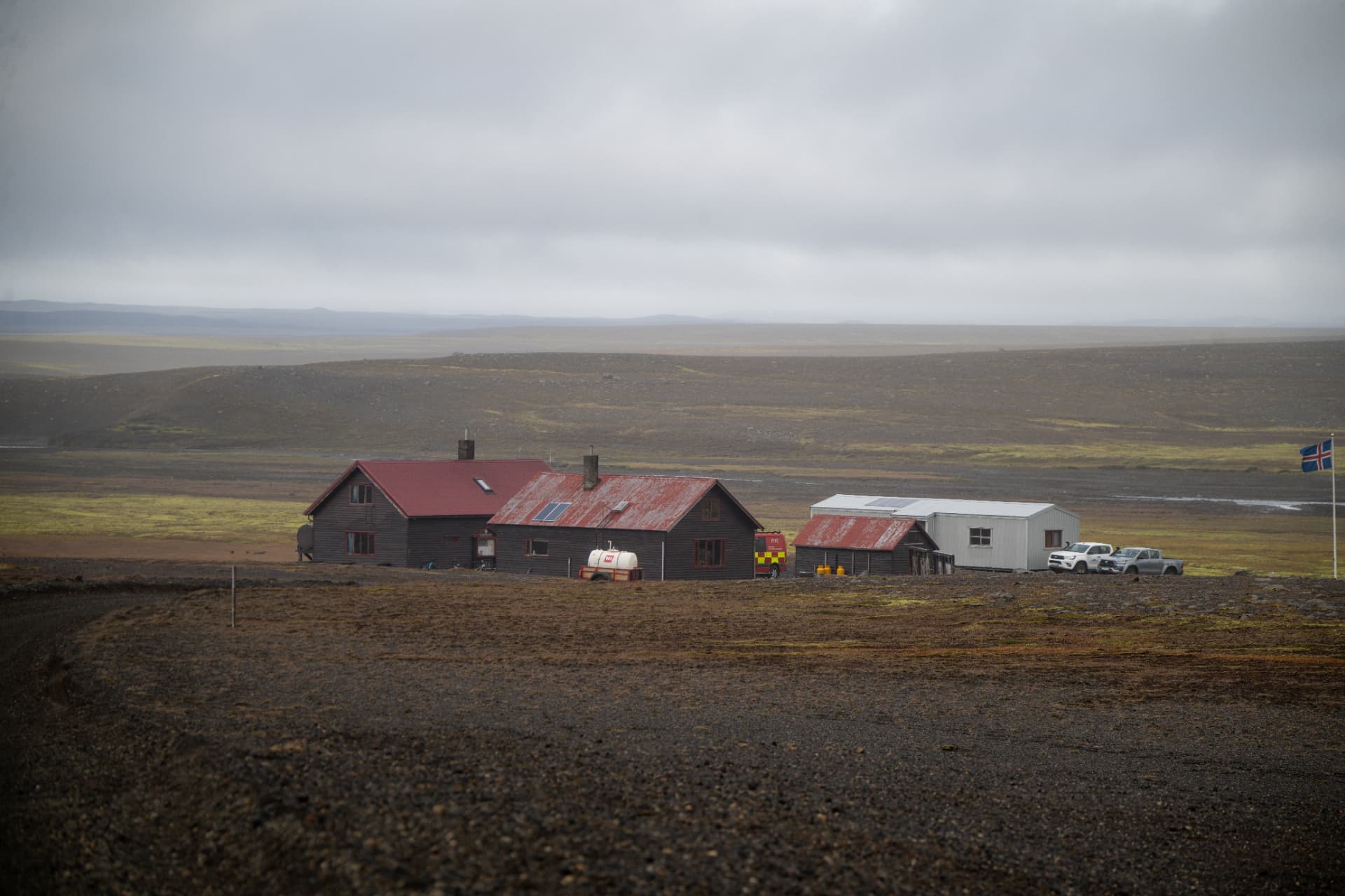 Remote highland farmstead with red roofs and an Icelandic flag under low grey cloud