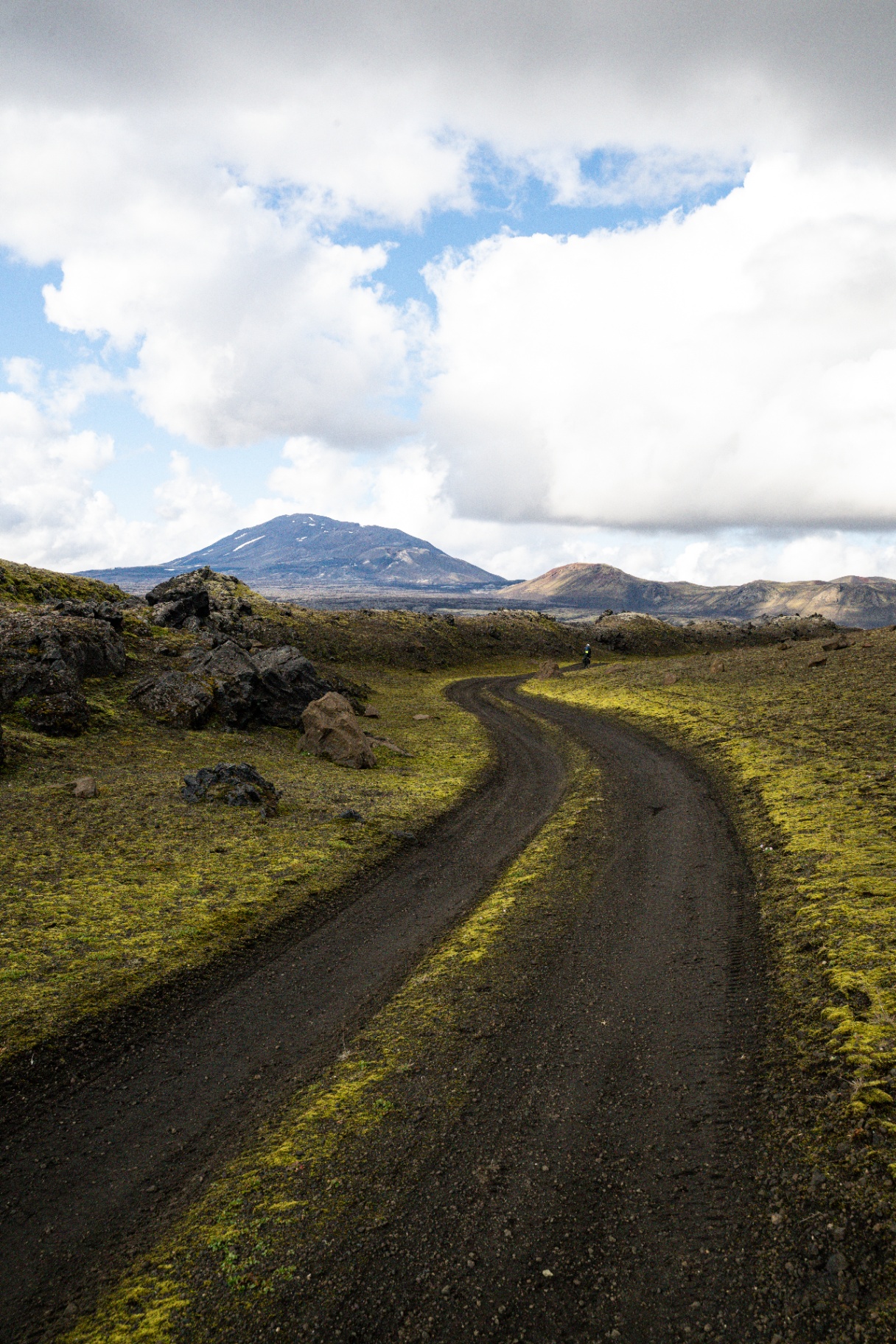 Gravel track curving through moss covered lava towards a distant snow streaked volcano