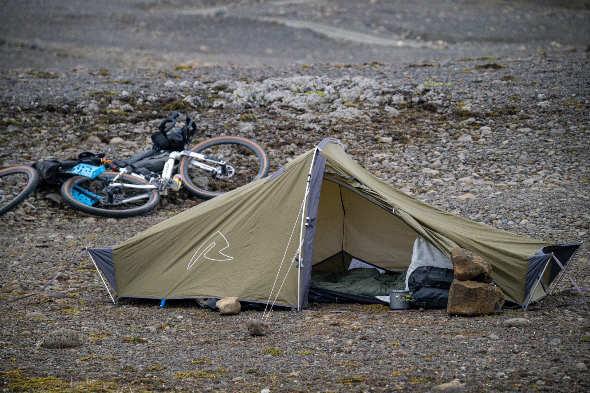 Olive bikepacking tent pitched on a stony riverbed beside a loaded gravel bike
