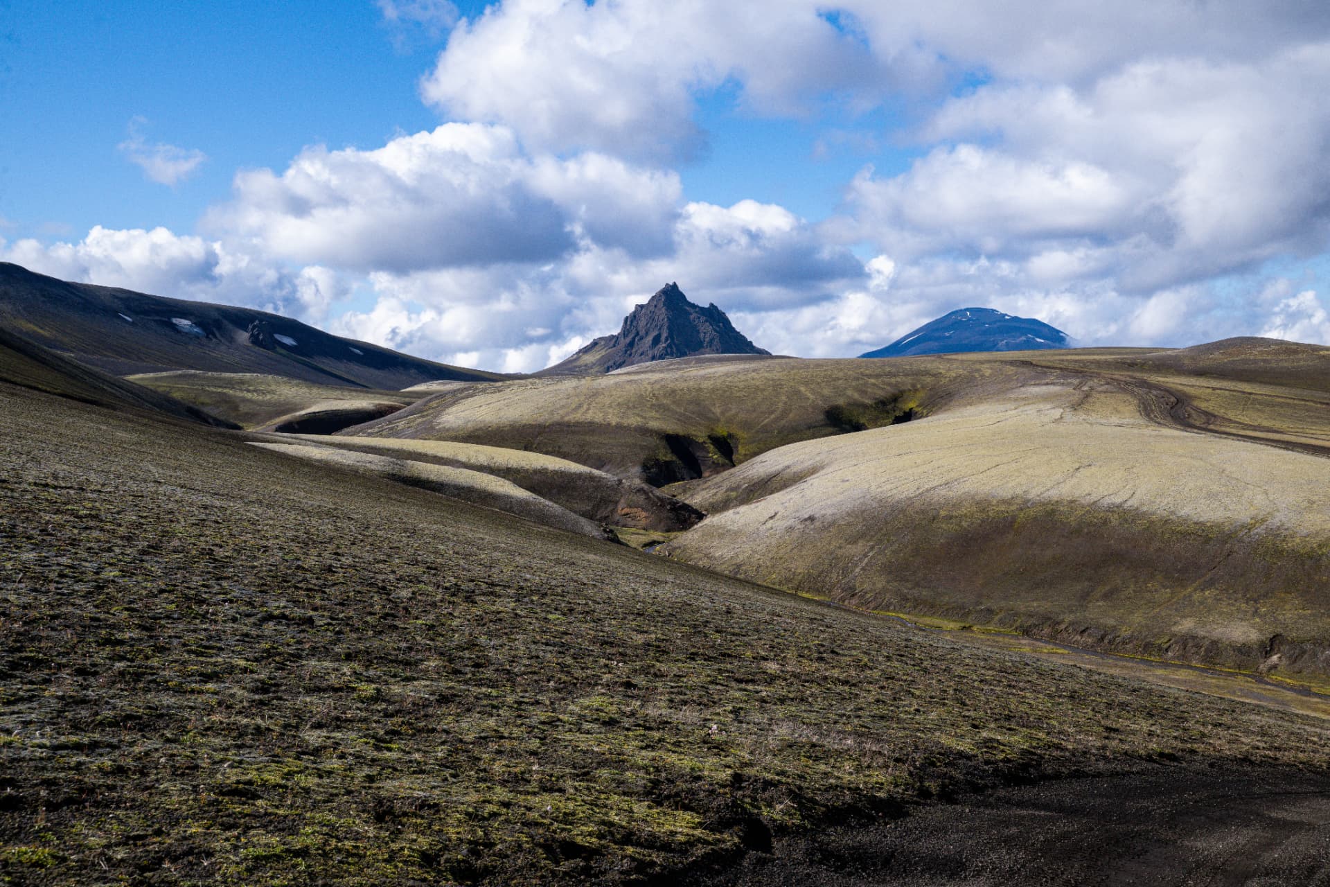 Jagged volcanic peak rising above rolling moss covered highland hills under cumulus cloud