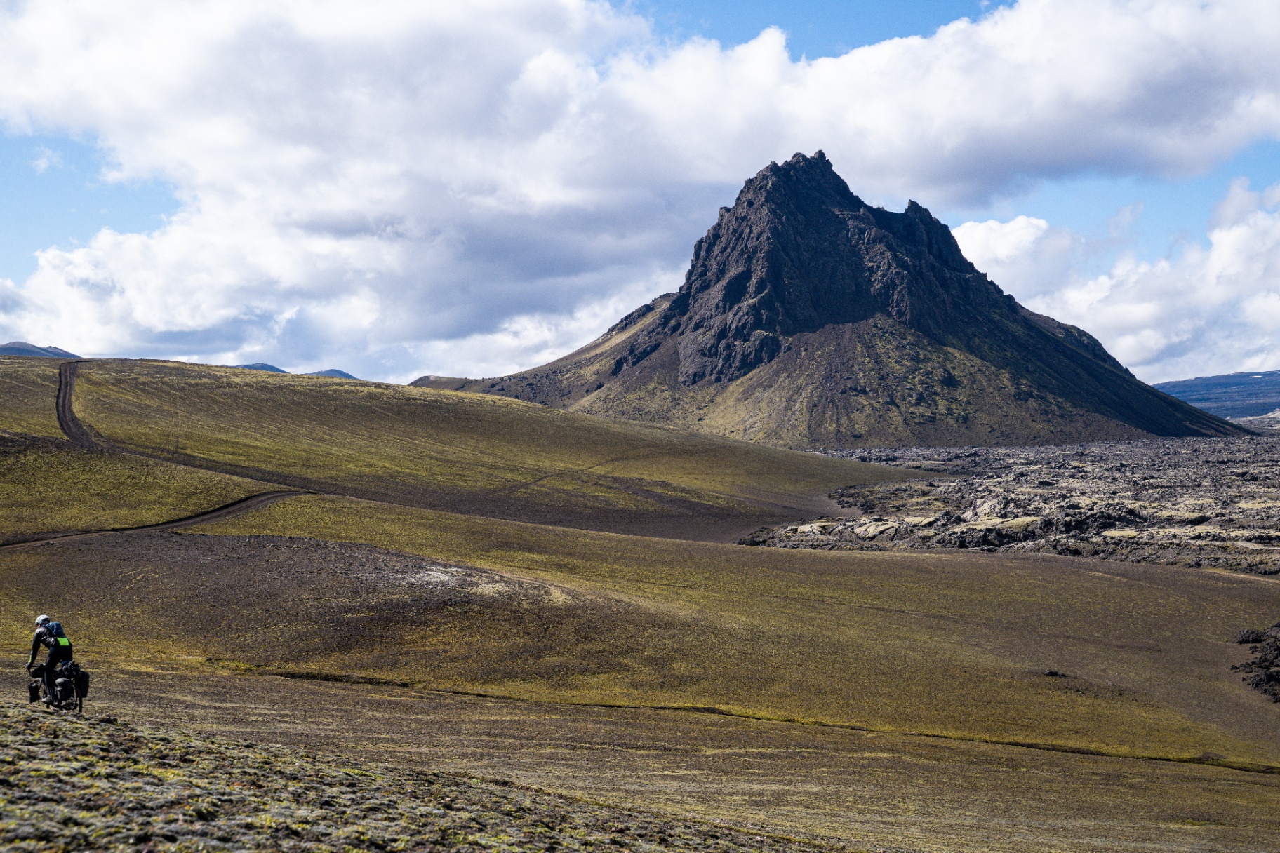 Loaded bikepacker crossing a vast mossy lava plain beneath a jagged volcanic peak