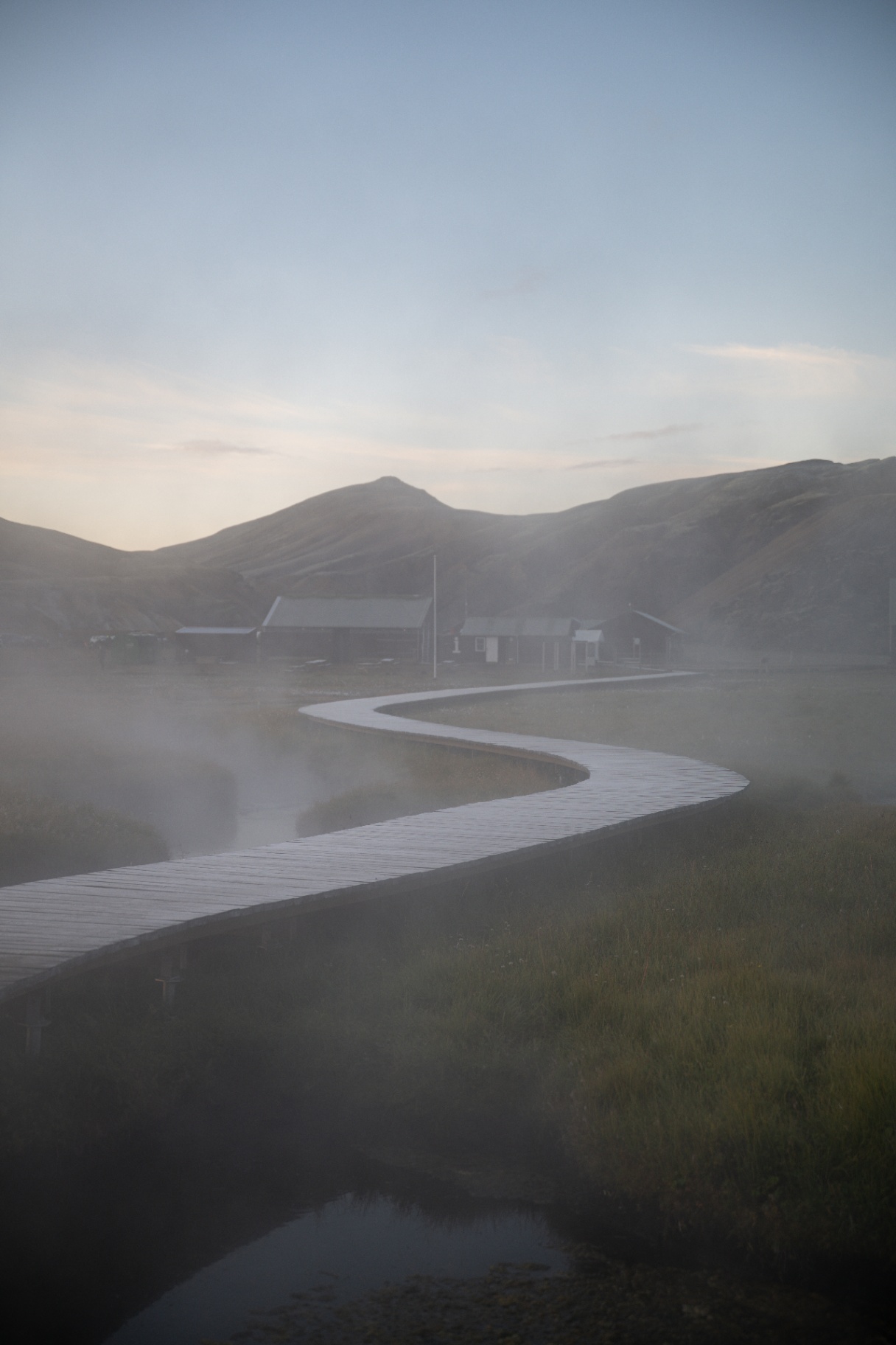 Boardwalk winding through steam and low light towards highland huts