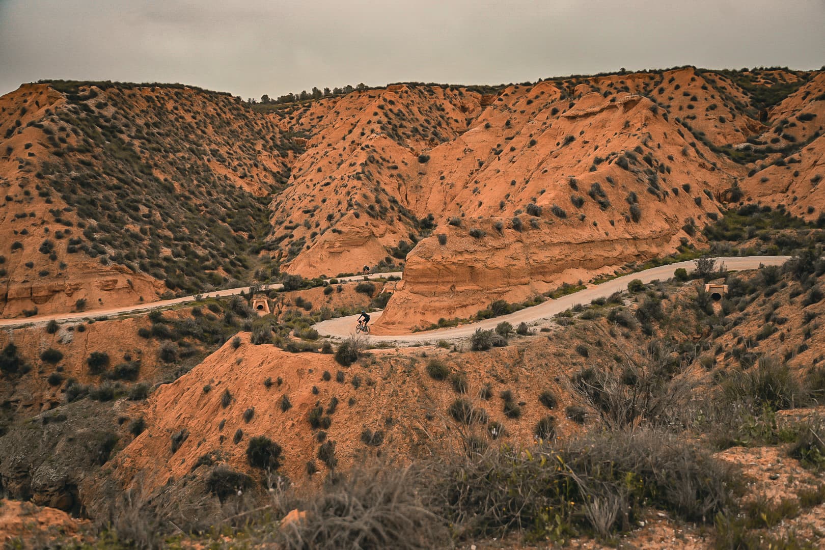 Two riders on a gravel road cut through orange badlands