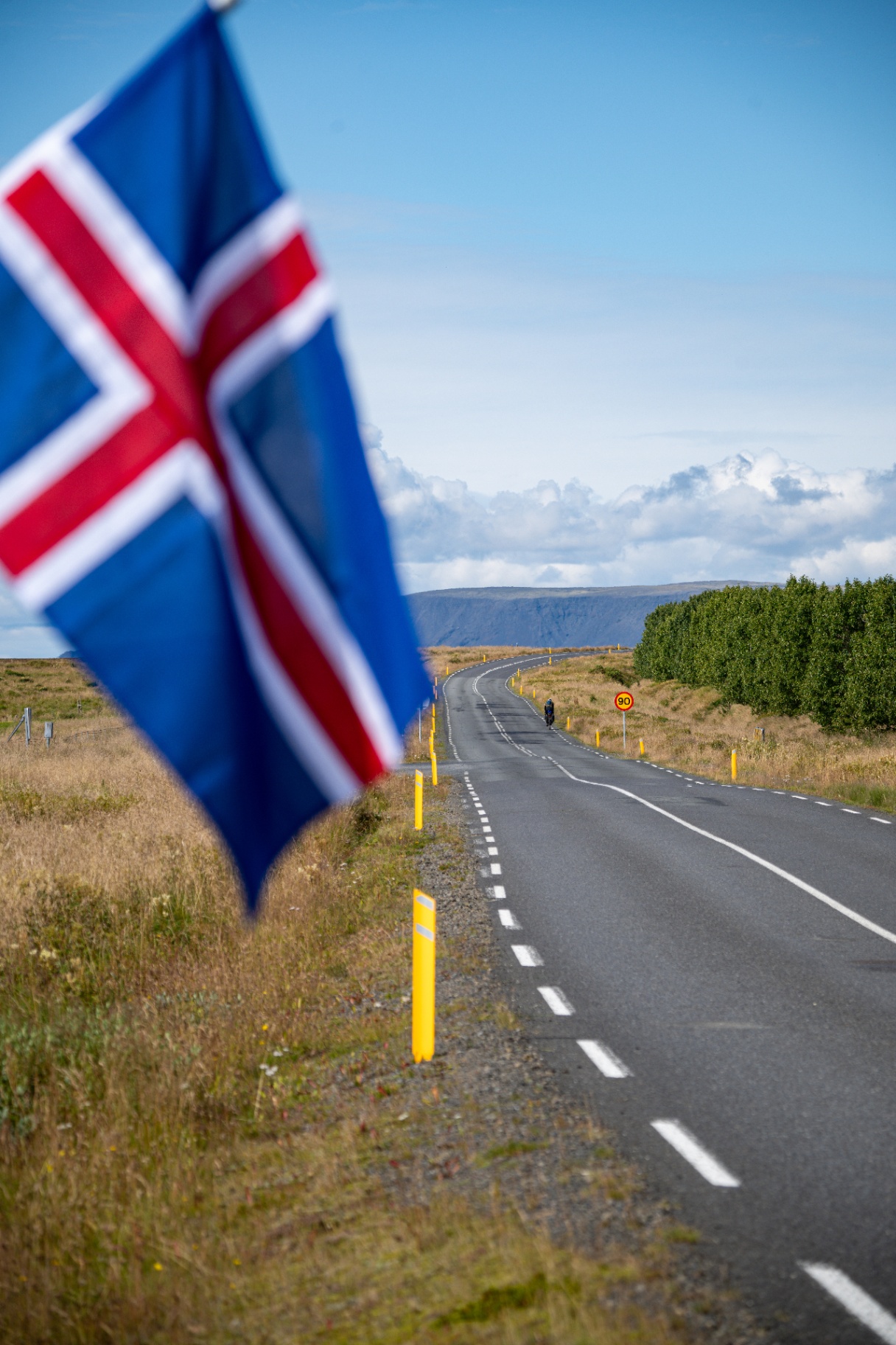 Icelandic flag in the foreground with a lone cyclist on the empty highway beyond