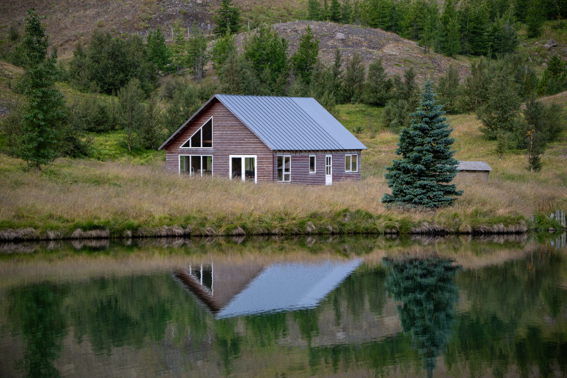 Timber cabin mirrored in a still lake below a wooded hillside